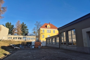 Dziedziniec między budynkami, z widocznym żółtym obiektem o czerwonym dachu w tle. Po bokach znajdują się niskie, szare zabudowania z oknami, a na środku placu leżą materiały budowlane. Nad całością rozciąga się czyste, niebieskie niebo.