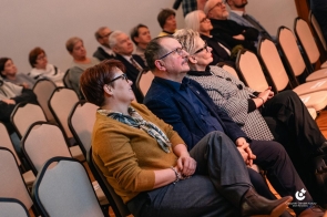 Publiczność siedzi na krzesłach w sali. W pierwszym rzędzie widoczne osoby w różnym wieku. Uczestnicy patrzą w kierunku sceny. W tle kolejne rzędy miejsc i ściana pomieszczenia.