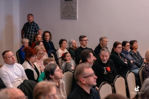 Publiczność siedzi na krzesłach w sali. W pierwszym planie widoczne osoby w różnym wieku, skierowane w stronę wydarzenia. W tle kolejne rzędy uczestników oraz ściana pomieszczenia.