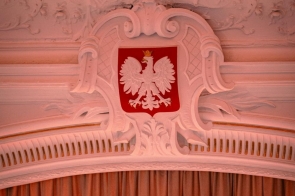 Godło Polski umieszczone centralnie na ścianie nad sceną. Poniżej kurtyna sceniczna. Wnętrze sali z dekoracyjnymi elementami architektonicznymi i oświetleniem w odcieniu czerwonym.
