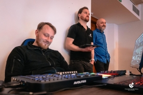 Trzech mężczyzn znajduje się przy stanowisku technicznym. Jeden siedzi przy konsoli dźwiękowej, dwóch stoi obok. Na stole widoczny sprzęt audio. W tle ściana pomieszczenia.