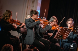 Muzycy siedzą na scenie i grają na instrumentach smyczkowych. Przed nimi pulpity z nutami. W tle widoczne elementy sceny oraz oświetlenie.