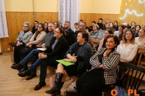 Grupa osób siedzi na krzesłach w sali. W tle widać kolejne rzędy uczestników, drewniane ściany, dekorację ze światełkami i gwiazdkami.