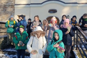 Grupa dzieci stoi na schodach przed budynkiem i pokazuje pomalowane kamienie. Dzieci są ubrane w zimowe kurtki, czapki i szaliki, a na ziemi widać śnieg.