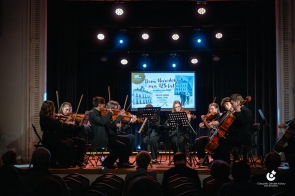 Zespół muzyków siedzi na scenie i gra na instrumentach. Nad nimi oświetlenie sceniczne. W tle ekran projekcyjny z wyświetloną grafiką. Przed sceną widoczna publiczność.