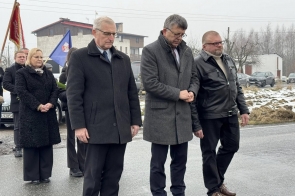 Grupa osób stoi na zewnątrz, część w mundurach, część w płaszczach. Jedna osoba oddaje salut. W tle widoczny pojazd oraz otoczenie z drzewami i zabudową.