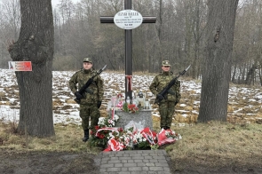 Dwóch żołnierzy w mundurach stoi przy pomniku w lesie. Przed nimi ułożone wieńce i zapalone znicze. W tle drzewa bez liści oraz leśne otoczenie.