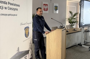 Mężczyzna stoi przy mównicy i przemawia. Obok roll-up z napisem oraz herbem. W tle ekran projekcyjny i fragment sali konferencyjnej z oknami oraz sprzętem.