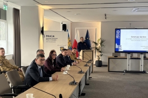 Uczestnicy siedzą przy stołach ustawionych w rzędzie w sali konferencyjnej. Na stołach mikrofony, dokumenty i butelki wody. Z przodu widoczny ekran prezentacyjny, w tle okna i ściany pomieszczenia.