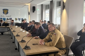 Uczestnicy siedzą przy długim stole konferencyjnym, pochyleni nad dokumentami. Na stole mikrofony, butelki wody i materiały. W tle okna wpuszczające światło dzienne oraz ściana sali.