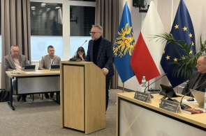 Kobieta stoi przy mównicy i przemawia do zgromadzonych. Obok widoczne flagi Polski i Unii Europejskiej. W tle siedzą radni powiatowi przy stole  z mikrofonami i dokumentami.