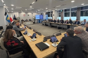 Sala sesyjna z ustawionymi w podkowę stołami i krzesłami.  Przy stołach siedzą obradujący radni powiatowi. Na stołach znajdują się mikrofony, dokumenty i butelki wody. W tle ekran prezentacyjny, flagi oraz okna wpuszczające światło dzienne.