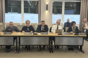 Zarząd Powiatu Cieszyńskiego siedzi przy długim stole  z mikrofonami i dokumentami. Przed nimi stoją butelki wody. W tle duże okna.