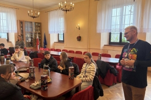 Grupa młodych osób siedzących przy stole z puszkami do zbiórki, słuchających stojącego obok mężczyzny, w jasnej sali z dużymi oknami i flagami w tle. WOŚP w Powiecie Cieszyńskim sztab w Zebrzydowicach.