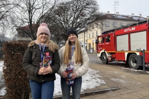 Dwie dziewczyny stojące na chodniku z puszkami do zbiórki, w tle widoczny czerwony wóz strażacki, budynki i zimowa sceneria. WOŚP w Powiecie Cieszyńskim.