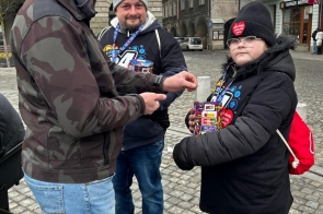 Mężczyzna wrzucający datek do puszki trzymanej przez dziecko w zimowym ubraniu, obok stoi drugi mężczyzna, a całość otoczona jest kolorowymi elementami graficznymi. Zbióka w Cieszynie.