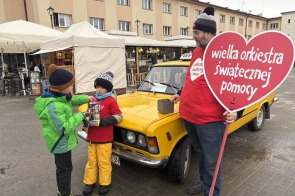 Dwóch chłopców stojących przy żółtym samochodzie wrzuca datki do puszki, obok mężczyzna trzyma duży czerwony znak w kształcie serca. WOŚP w Powiecie Cieszyńskim kwestujący w Wiśle.
