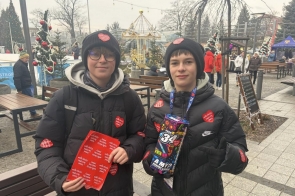 Dwóch chłopców w zimowych kurtkach i czapkach trzyma puszkę do zbiórki oraz naklejki w kształcie serc, stojąc wśród świątecznych dekoracji i stoisk. WOŚP w Powiecie Cieszyńskim.