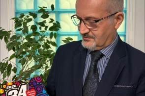 Wicestrosta Cieszyński Mieczysław Szczurek w garniturze trzyma i ogląda dokument związany z finałem Wielkiej Orkiestry Świątecznej Pomocy, stojąc na tle rośliny i przeszklonej ściany.