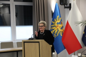 Beata Macura Członkini Zarządu stoi przy mównicy z mikrofonem, ubrana na czarno, w okularach. Obok widoczne są flagi, w tle zasłona oraz fragment sali sesyjnej.