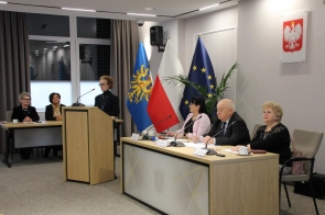 Klaudia Bohucka Rzecznik Konsumentów ubrana w czarna marynarkę stoi  przy mównicy, a obok, przy stole z mikrofonami, siedzi kilka osób uczestniczących. W tle widoczne są flagi Polski i Unii Europejskiej oraz godło państwowe.