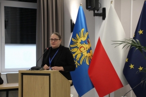 Agnieszka Baszczyńska Dyrektor PCPRu stoi przy mównicy z mikrofonem, ubrana w ciemny strój i okulary, z identyfikatorem na szyi. Obok widoczne są flagi oraz fragment rośliny, w tle zasłona i ściana sali.