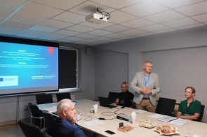 Sala z długim stołem i ekranem projekcyjnym. Kilka osób siedzi przy stole, jedna osoba stoi i ma złożone ręce. Na stole znajdują się laptopy, dokumenty oraz filiżanki. Pod  sufitem zamontowany jest projektor.