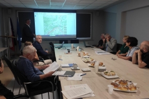 Sala z długim stołem i ekranem projekcyjnym wyświetlającym mapę. Kilka osób siedzi po obu stronach stołu, jedna osoba stoi obok ekranu. W tle widoczne flagi.