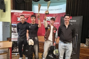 Czterech młodych mężczyzn stojących na scenie, dwóch z nich unosi puchary, a pozostali trzymają torby z nagrodami; w tle banery szkoły.