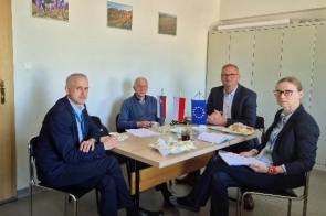 Cztery osoby siedzą wokół stołu, na których znajdują się dokumenty oraz filiżanki. Na stole ustawione są małe flagi Słowacji, Unii Europejskiej i Polski. W tle widoczna jest jasna ściana z obrazami oraz szafa.