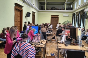 W dużej sali wiele osób pracuje przy stanowiskach fryzjerskich, strzygąc klientów siedzących na fotelach; wokół widoczne stoły z narzędziami i przewody rozciągnięte po podłodze.
