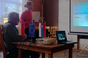 Kobieta stojąca przy stole trzyma dokument. Na stole znajduje się projektor, laptop oraz małe flagi Polski, Unii Europejskiej i Słowacji. Obok ustawiony jest ekran z wyświetloną prezentacją programu Interreg Polska-Słowacja. 