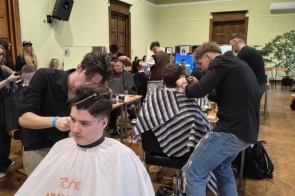 Kilku fryzjerów pracujących jednocześnie przy klientach siedzących na krzesłach, w dużej sali z wysokim sufitem i stanowiskami ustawionymi w rzędach.