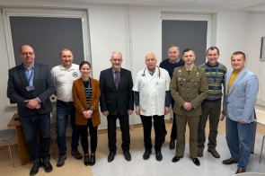 Grupa dziewięciu osób ustawionych w rzędzie w jasnym pomieszczeniu; wśród nich mężczyzna w mundurze oraz osoba w białym kitlu.