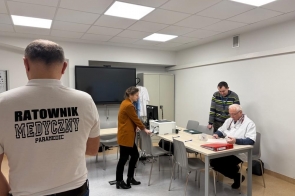 Kilka osób w jasnej sali: mężczyzna w koszulce z napisem „Ratownik Medyczny”, kobieta przesuwająca krzesło oraz dwóch mężczyzn przy stole, z których jeden przegląda dokumenty.
