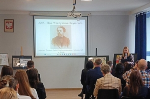 Osoby siedzą na krzesłach i słuchają wystąpienia. Z przodu stoi kobieta z mikrofonem. Na ekranie wyświetlona jest prezentacja z Władysławem Reymontem. 