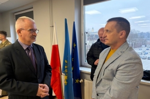Dwóch mężczyzn prowadzących rozmowę w pomieszczeniu biurowym,po lewej stoi Mieczysław Szczurek Wicestarosta Cieszyński, obok flag Polski, Unii Europejskiej i regionu; w tle widoczne okno z zimowym krajobrazem.