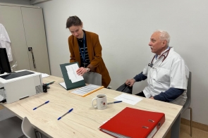 Kobieta stojąca przy stole przegląda dokumenty w teczce, obok siedzący starszy mężczyzna ze stetoskopem obserwuje sytuację; na blacie widoczne segregatory, długopisy i kubek.