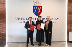 Trzy osoby stojące przed jasną ścianą z herbem. Dwie osoby trzymają czerwone teczki, jedna osoba trzyma certyfikat. Wszyscy ubrani są w formalne stroje. Widoczna jest podłoga z jasnych płytek oraz fragment ceglanej ściany po bokach.