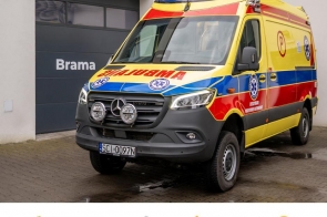 Ambulans ratownictwa medycznego zaparkowany na placu, w tle widoczne zabudowania