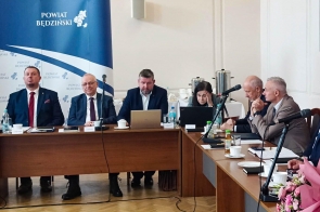 Przy stołach ustawionych w kształcie litery U siedzą uczestnicy spotkania z laptopami, dokumentami i mikrofonami. W tle widoczny jest baner z napisem „Powiat Będziński”.