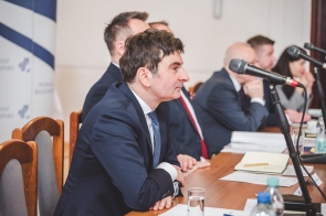 Przy stole siedzą uczestnicy spotkania w garniturach, przed nimi stoją mikrofony, dokumenty, notesy i szklanki z wodą. Na pierwszym planie widoczny jest Wojewoda Śląski Marek Wójcik mężczyzna w granatowym garniturze siedzący bokiem do stołu.
