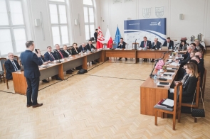 W jasnej sali przy stołach ustawionych w kształcie litery U siedzą uczestnicy spotkania z mikrofonami, dokumentami i laptopami. Na środku stoi mężczyzna mówiący do mikrofonu, a w tle widoczne są flagi Polski i Unii Europejskiej oraz baner z napisem „Powiat Będziński”.