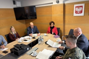 W sali konferencyjnej kilka osób siedzi przy stole i prowadzi rozmowę. Na ścianie widoczny jest biały orzeł na czerwonym tle oraz zamontowany telewizor. Na stole leżą dokumenty, notesy i telefony, a przed uczestnikami stoją filiżanki z kawą i butelki wody.