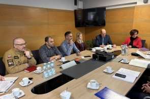 W sali konferencyjnej kilka osób siedzi przy dużym stole podczas spotkania. Na stole stoją filiżanki z kawą, butelki wody oraz leżą dokumenty i telefony. Uczestnicy, w tym osoba w mundurze, słuchają i przeglądają materiały.