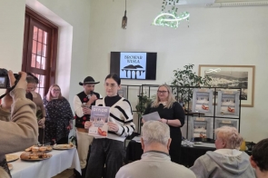 W sali kilka osób siedzi przodem do wydarzenia. Na środku stoi młoda kobieta trzymająca plakat z napisem „Dyplom”, obok niej znajdują się inne osoby biorące udział w uroczystości. Po lewej stronie ktoś robi zdjęcie aparatem.