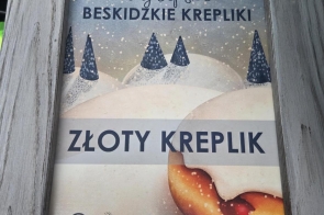 W drewnianej ramce znajduje się dyplom za I miejsce w konkursie „Najlepsze Beskidzkie Krepliki – Złoty Kreplik”. Na dole widoczny jest fragment pączka, a przed ramką leży złota łyżka.