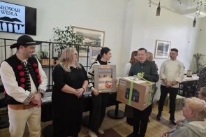 W jasnym pomieszczeniu kilka osób stoi obok siebie podczas spotkania. Jeden z mężczyzn trzyma duże pudełko przewiązane zieloną wstążką i pokazuje je zgromadzonym.
