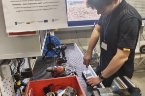 Młody mężczyzna z identyfikatorem na szyi pracuje przy stanowisku warsztatowym, używając śrubokręta do rozkręcania metalowego elementu zamocowanego w imadle. Na stole znajdują się narzędzia, wiertarka, przewody oraz czerwony pojemnik z częściami oznaczony napisem „Wadliwe części”. W tle widać monitor i tablicę informacyjną.