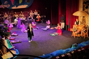 Scena teatralna z wieloma osobami i rekwizytami; w tle gra orkiestra, a na pierwszym planie aktorzy stoją i klęczą wśród porozkładanych walizek oraz elementów scenografii.
